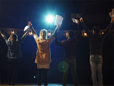 A group of people standing outdoors at night with their arms raised in celebration, illuminated by bright stage lights.