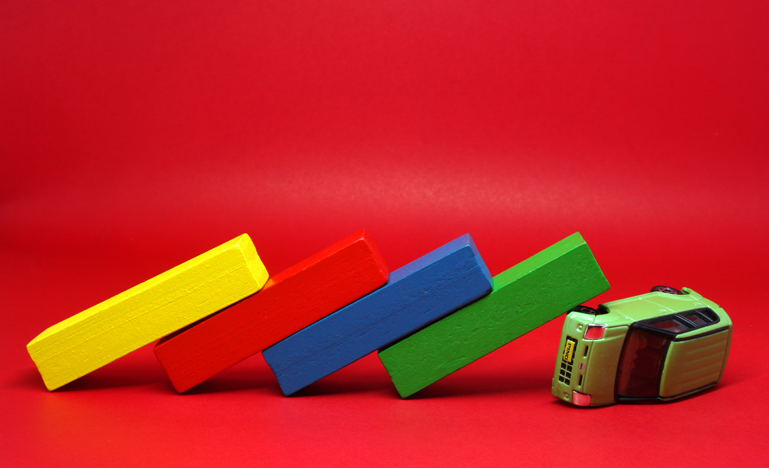 A toy bus is tipped over with four colored blocks—yellow, red, blue, and green—stacked and falling like dominoes against it, set on a red background.