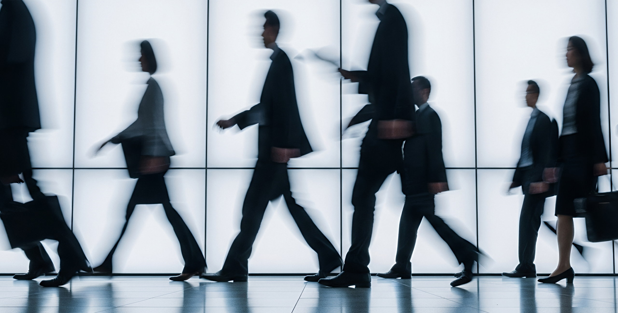 Blurred silhouettes of business professionals walking past a glass wall, suggesting movement and anonymity