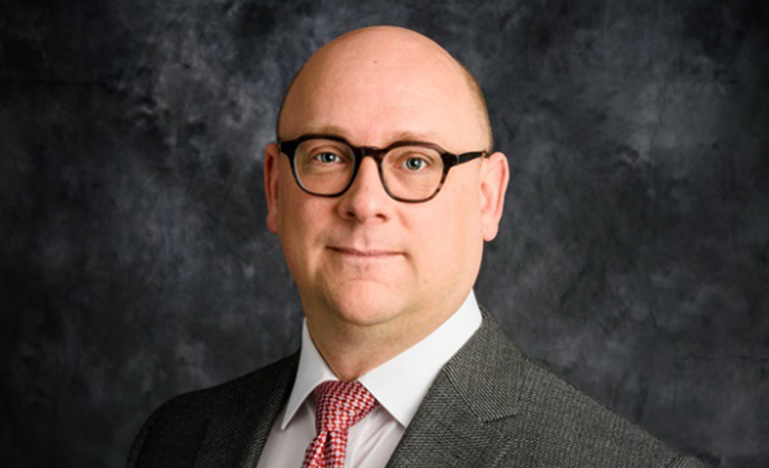 David Koski, a white man wearing glasses and a gray suit jacket, poses in front of a dark background