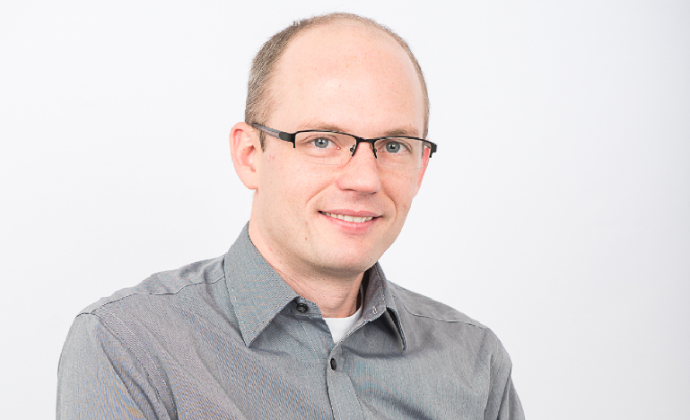 Trevor Tombe, a white man wearing glasses and a gray collared shirt, poses against a white backdrop