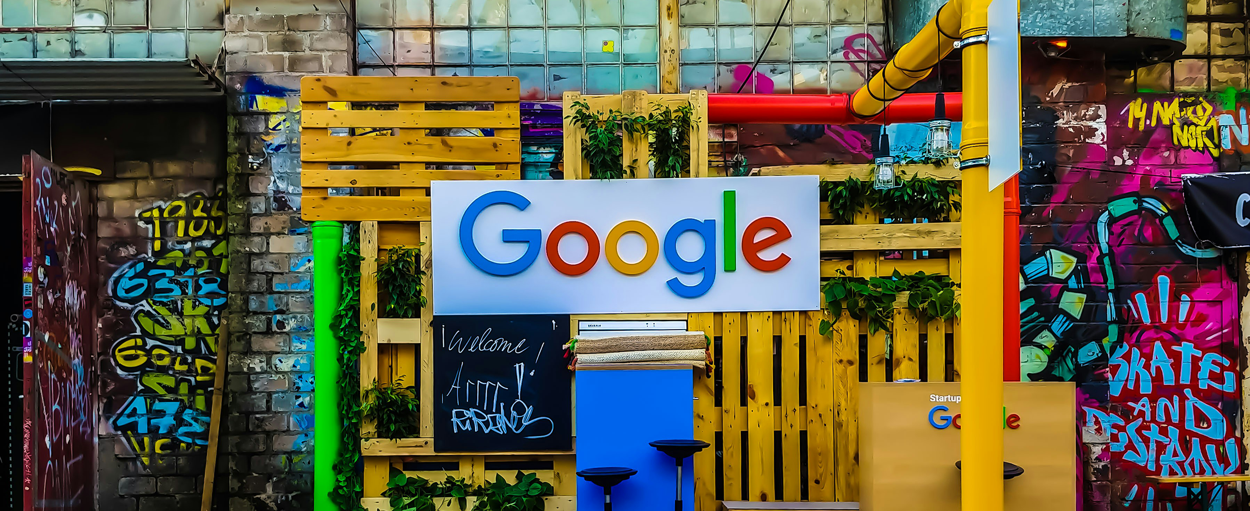 A decorated area with a large Google sign, wooden pallets, green plants, and graffiti-covered walls in the background.