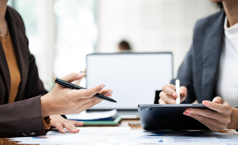 Two people in business attire discussing documents; one holds a pen and paper, the other uses a stylus on a tablet. A laptop is open on the desk in the background.