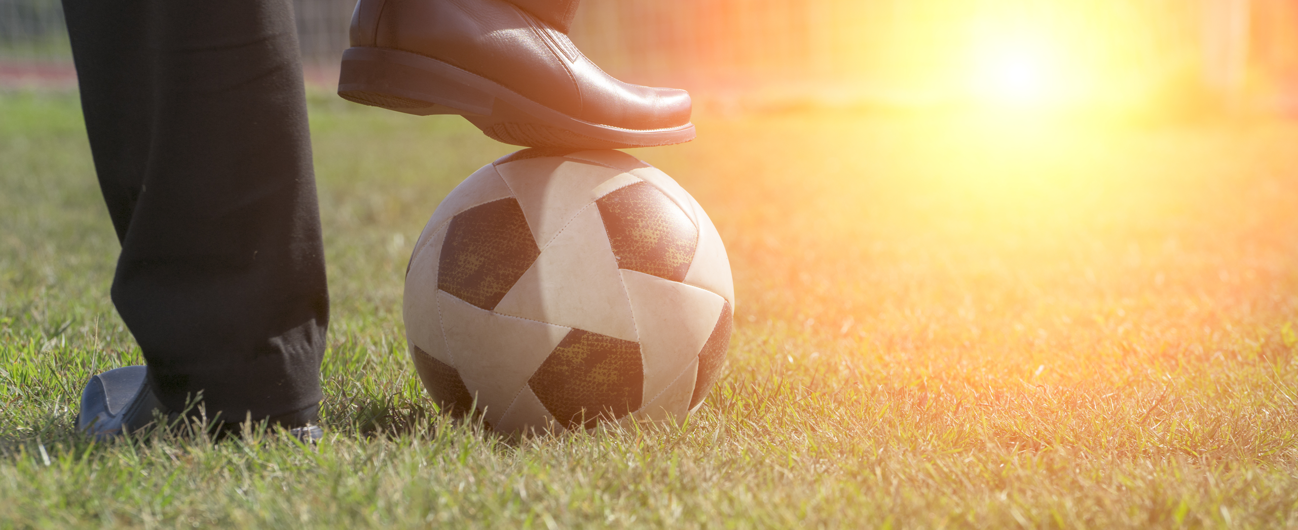 close-up of a person’s foot resting on a soccer ball on a grassy field, with sunlight in the background.