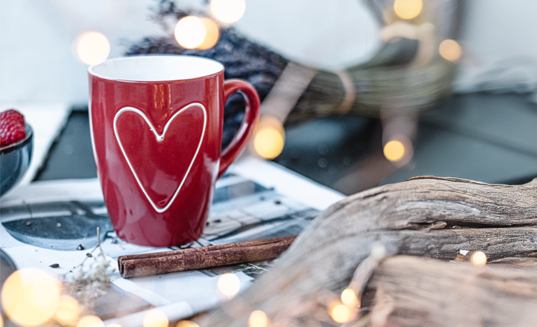 A red mug with a heart design sits on a table among Christmas lights and other festive decor
