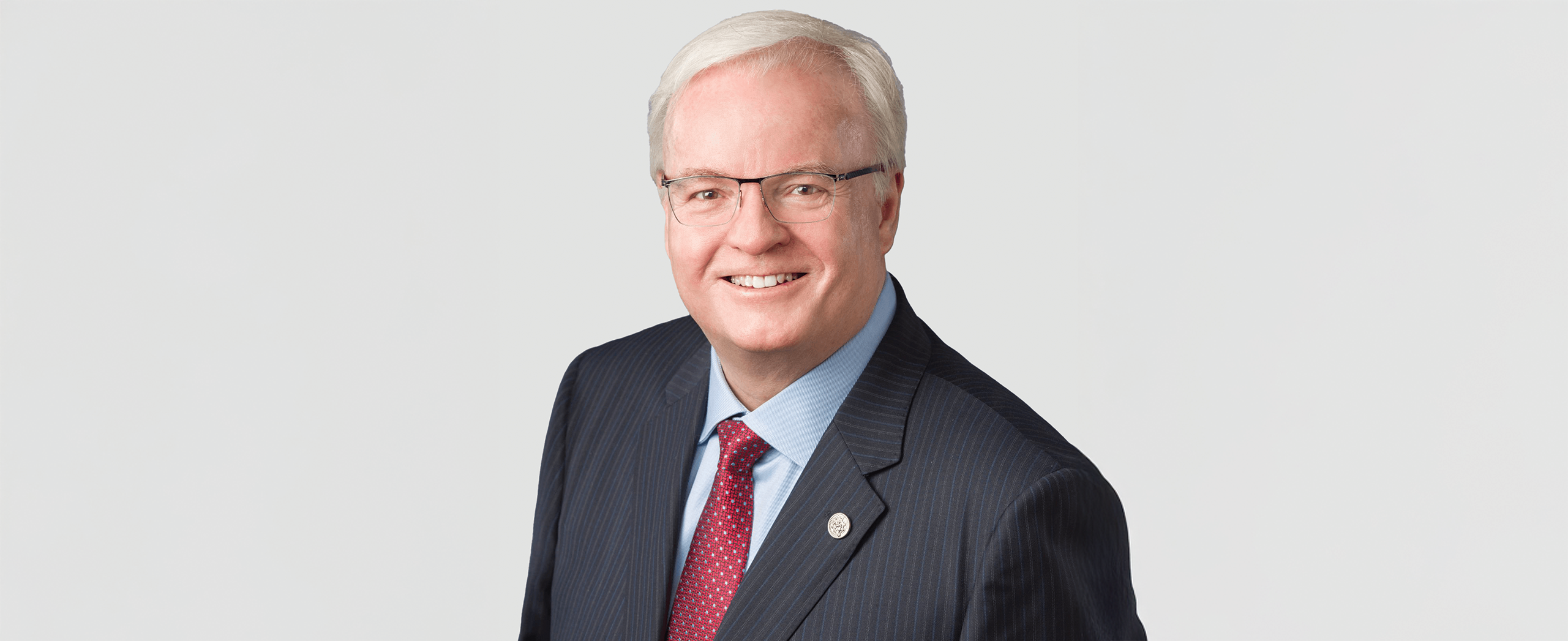 Tracey Stock, and man with short white hair and glasses wearing a dark pinstripe suit, light blue shirt, and red patterned tie, poses against a plain light background.
