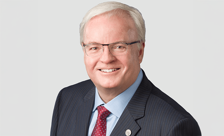 Tracey Stock, and man with short white hair and glasses wearing a dark pinstripe suit, light blue shirt, and red patterned tie, poses against a plain light background.