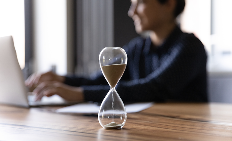 A professional woman in a suit working on a laptop, with an hourglass in the foreground.