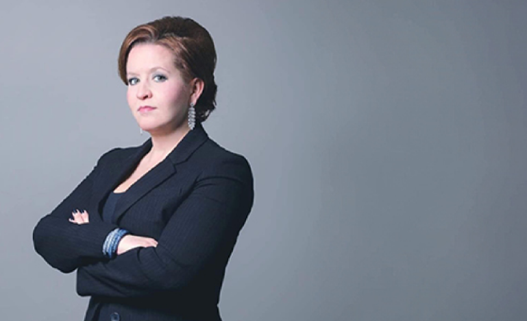 Kathy Briere, a woman with short brown hair and wearing a dark suit jacket, poses in front of a gray background with her arms crossed