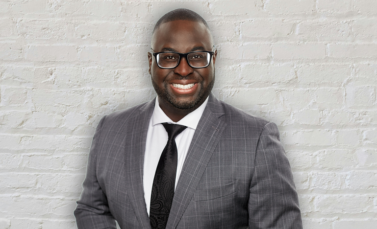 Mosi Taylor-Cole, a Black man with short hair and glasses, poses in front of a white brick wall wearing a grey suit.